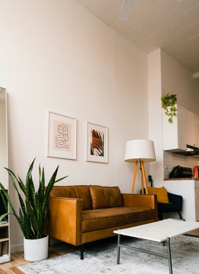 Modern living room with a caramel leather sofa, two minimalist framed artworks on a white wall, a tall snake plant in a white pot, and a marble-top coffee table on a light rug. A wooden tripod floor lamp stands beside the sofa, with a small open kitchen visible in the background featuring white cabinets and a trailing green plant.