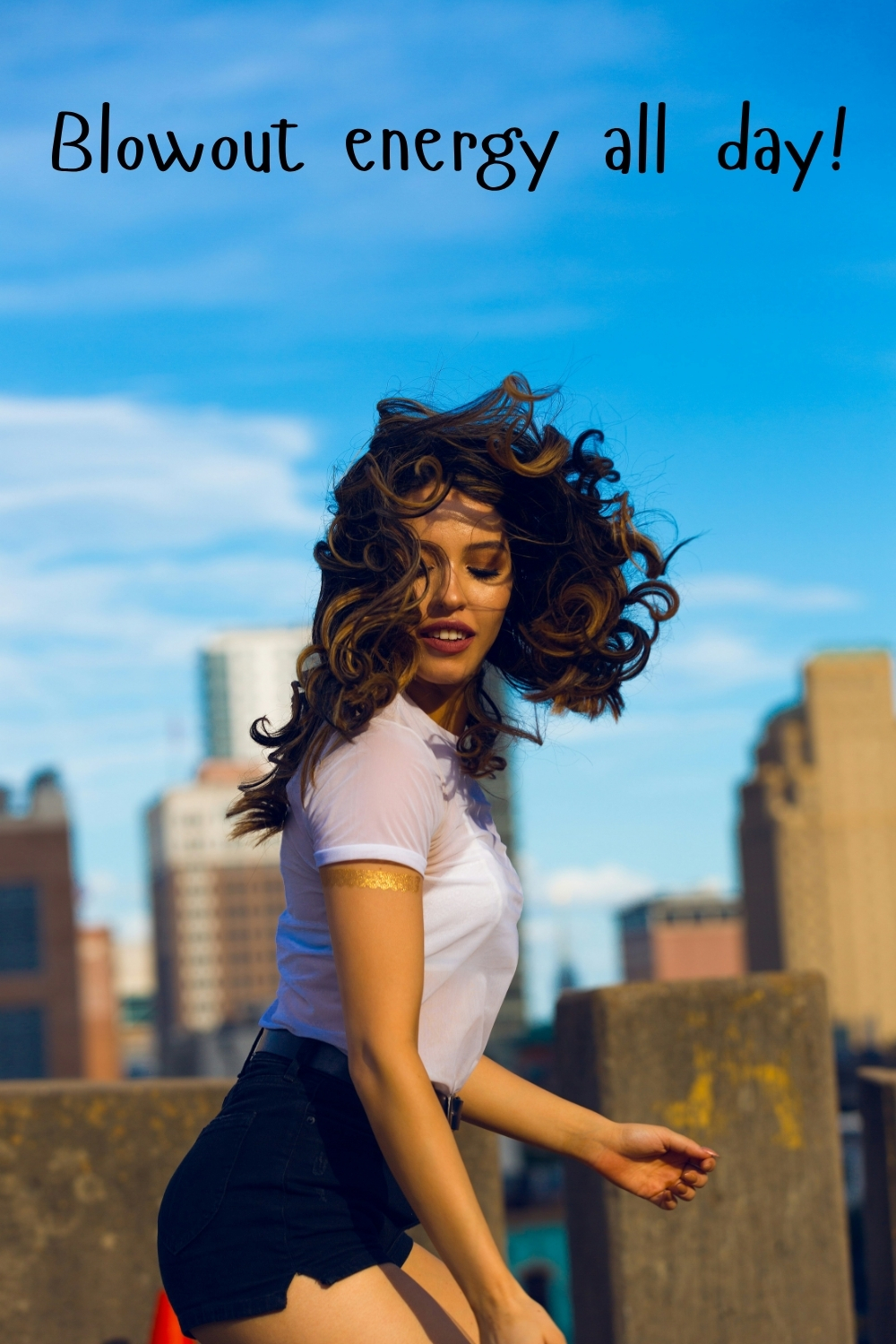 Woman with voluminous curly hair moving in the wind on a rooftop with city skyline and the quote “Blowout energy all day!”