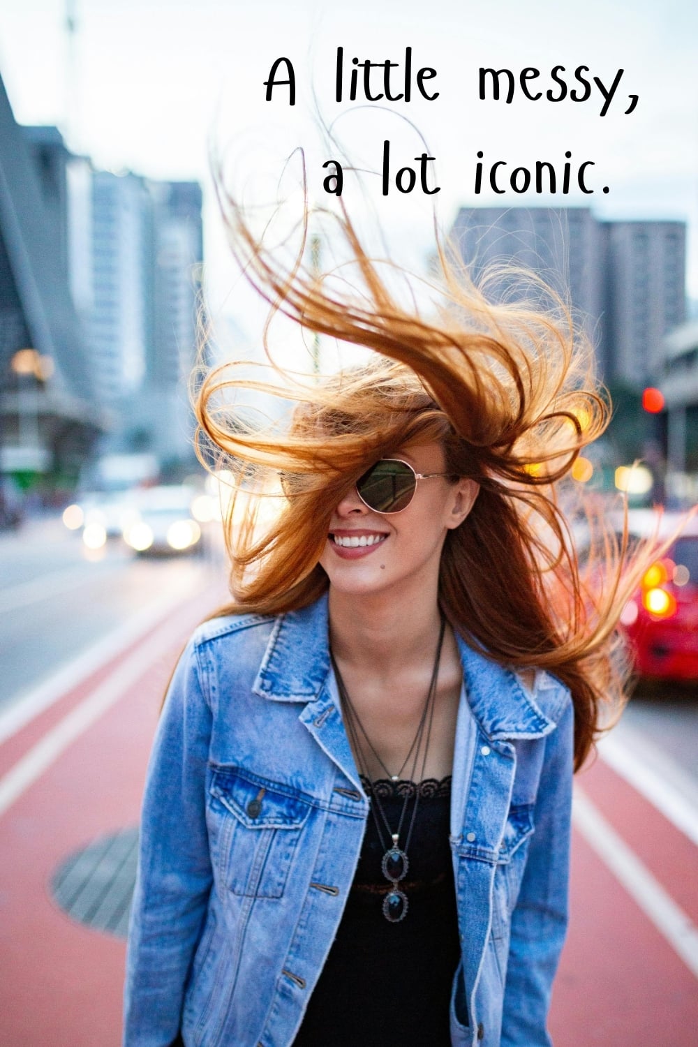 Smiling woman with flowing red hair wearing sunglasses and a denim jacket in a city setting with the quote “A little messy, a lot iconic.”