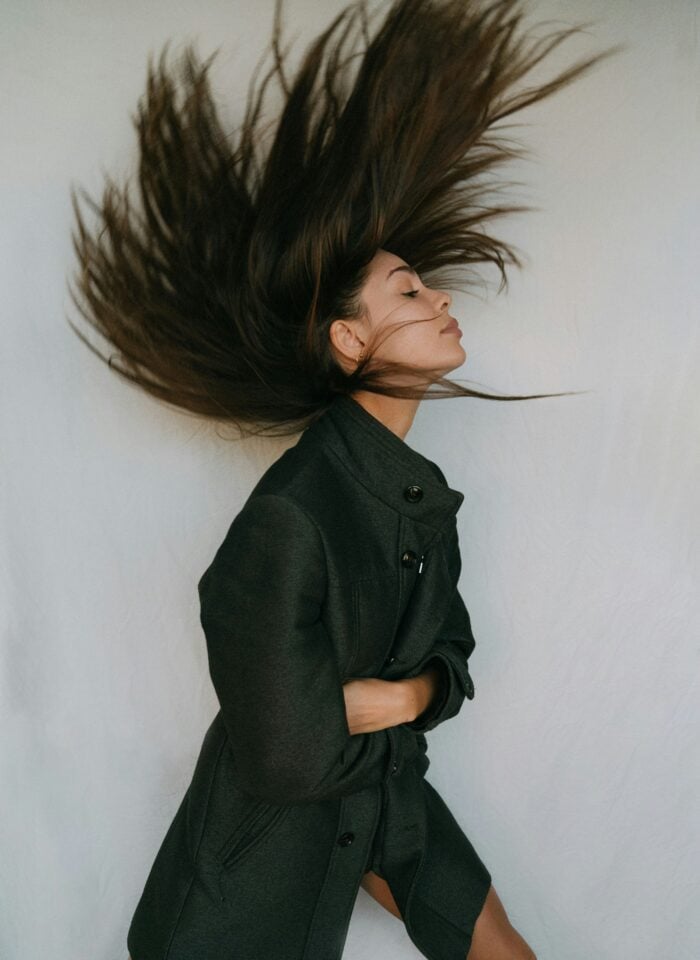 Woman flipping long dark hair mid-motion wearing a black coat against a plain backdrop