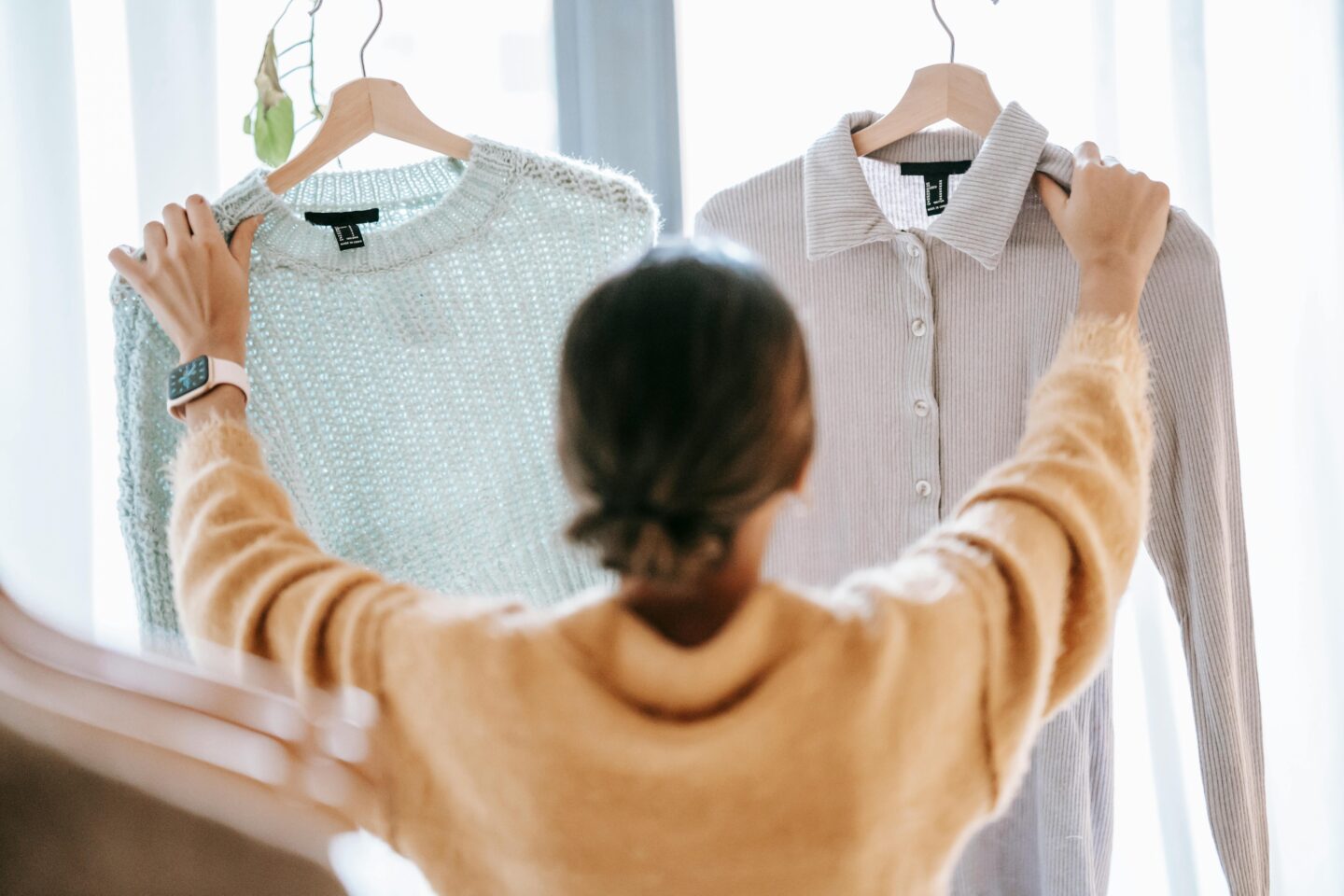 Woman choosing between neutral knit tops on hangers while planning versatile everyday outfits and casual wardrobe combinations.