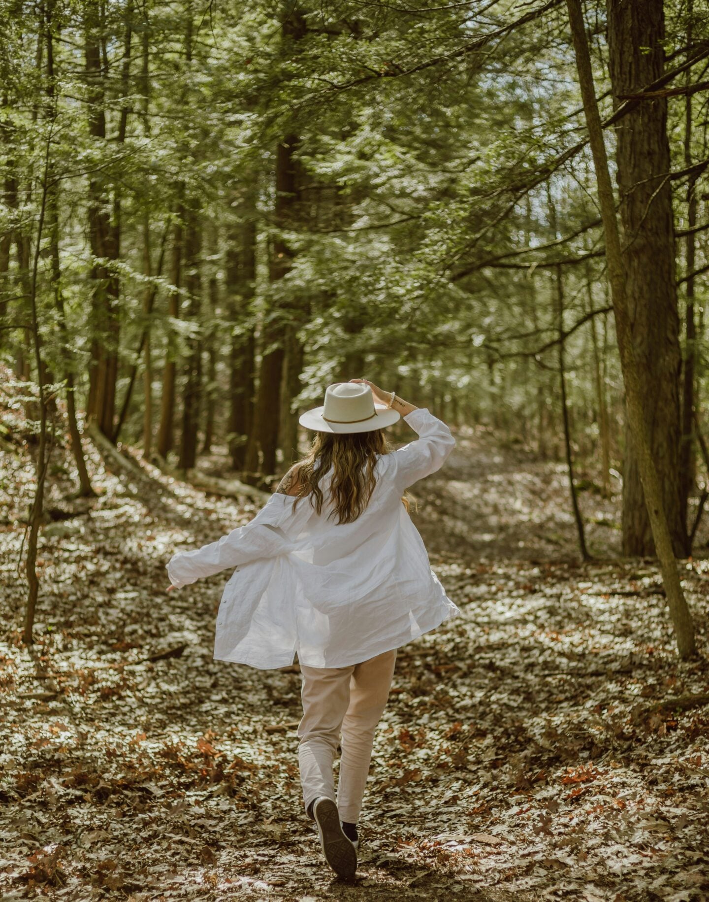 Woman wearing a white shirt and wide-brim hat walking through a sunlit forest surrounded by tall trees and fallen leaves.