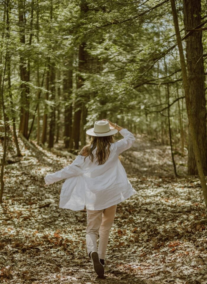 Woman wearing a white shirt and wide-brim hat walking through a sunlit forest surrounded by tall trees and fallen leaves.