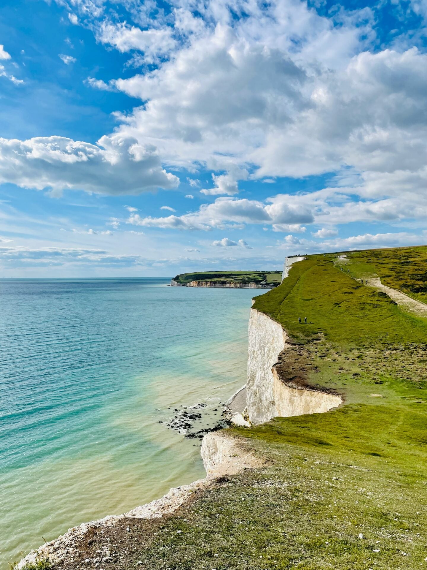 White chalk cliffs overlooking the turquoise sea at Beachy Head, with a grassy coastal path stretching along the cliff tops beneath a dramatic blue sky filled with large white clouds.