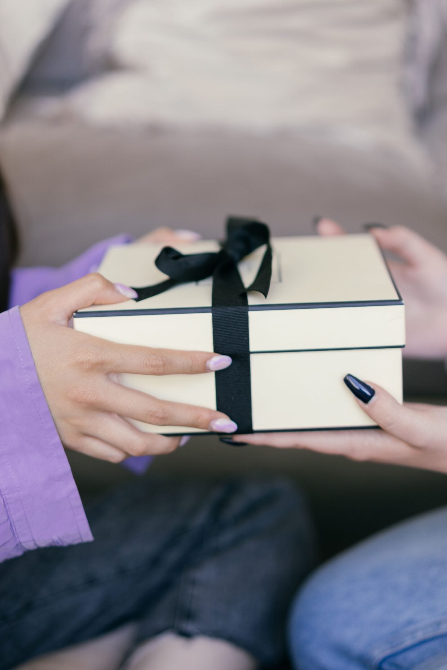 Two women exchanging a gift box wrapped with a black ribbon, with one woman handing the present to the other. The close-up image captures a thoughtful gift-giving moment.