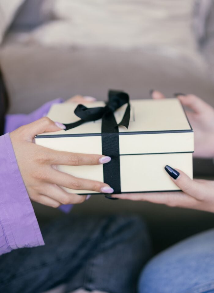 Two women exchanging a gift box wrapped with a black ribbon, with one woman handing the present to the other. The close-up image captures a thoughtful gift-giving moment.
