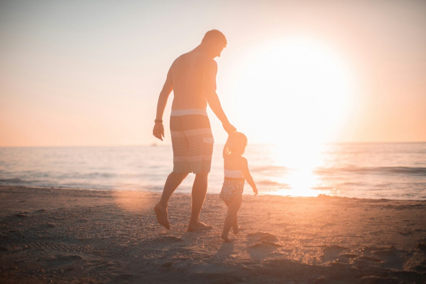 Father and young child walking together along a beach at sunset.