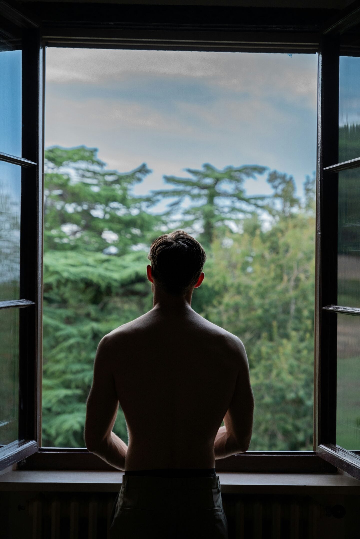 Man standing at an open window looking out over trees and the surrounding landscape.
