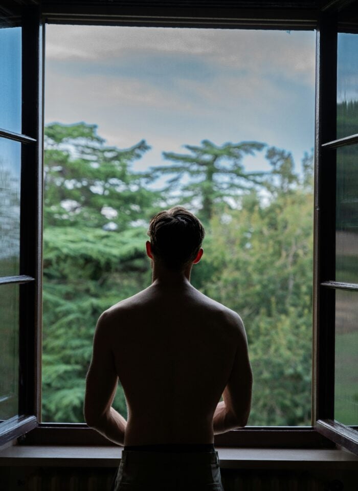 Man standing at an open window looking out over trees and the surrounding landscape.