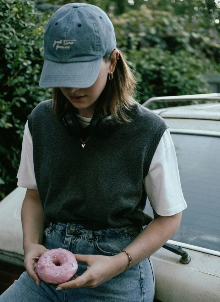 Person wearing a blue baseball cap, grey sweater vest over a white T-shirt, and jeans while holding a pink frosted doughnut, leaning against a vintage car outdoors.