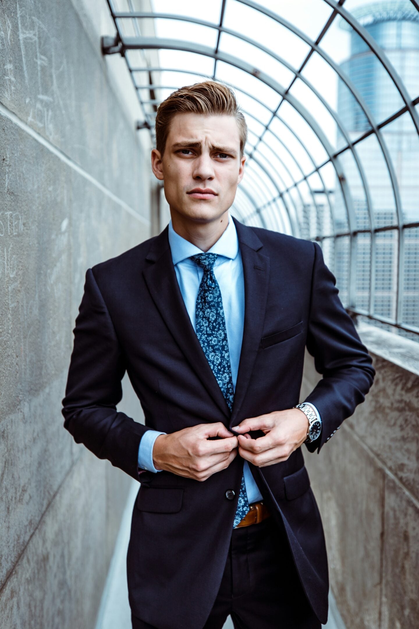 Young professional adjusting a navy suit jacket while standing in a modern urban walkway, demonstrating polished business style with personality.