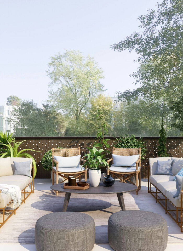 Modern outdoor living space with two cream-colored sofas, woven accent chairs, gray poufs, and a round coffee table arranged on a spacious terrace surrounded by lush greenery. Potted plants, decorative cushions, and warm natural materials create a comfortable, home-like atmosphere with peaceful garden views.