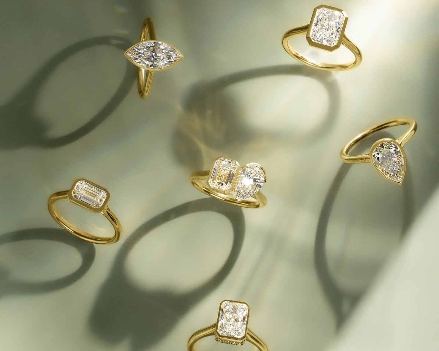 Collection of elegant gold rings featuring marquise, emerald, pear, and radiant-cut diamonds displayed against a soft green background with artistic shadows.
