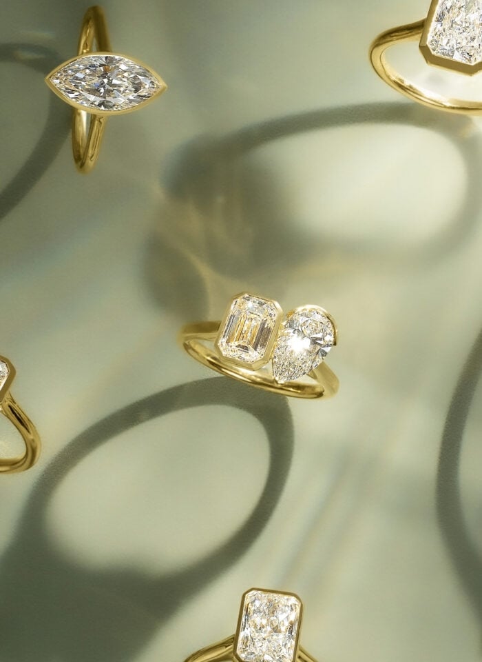 Collection of elegant gold rings featuring marquise, emerald, pear, and radiant-cut diamonds displayed against a soft green background with artistic shadows.