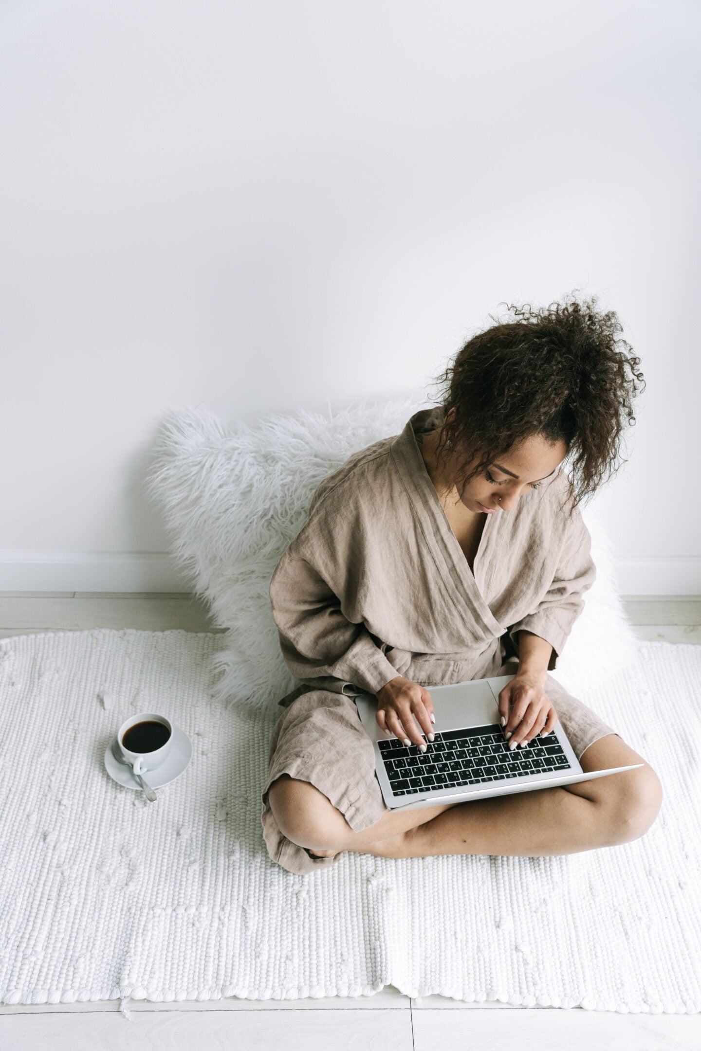 Woman wearing neutral beige loungewear while sitting on a white rug and working on a laptop at home, with a cup of coffee nearby in a bright, minimalist living space.
