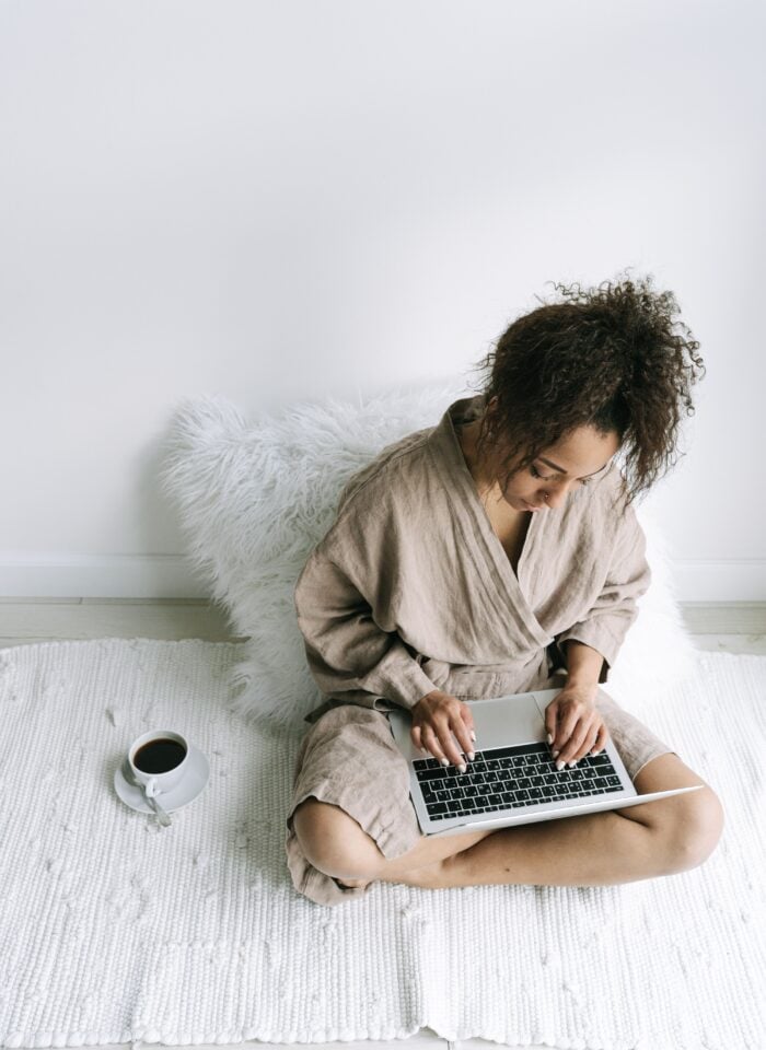 Woman wearing neutral beige loungewear while sitting on a white rug and working on a laptop at home, with a cup of coffee nearby in a bright, minimalist living space.