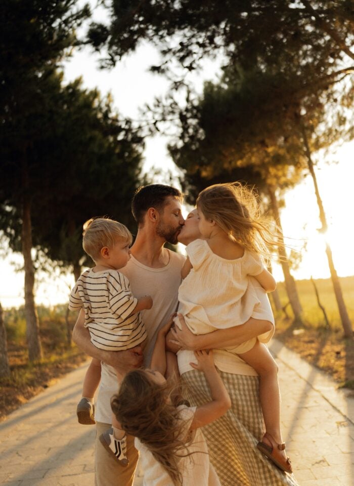 Parents sharing a kiss while holding their children during a sunset walk.