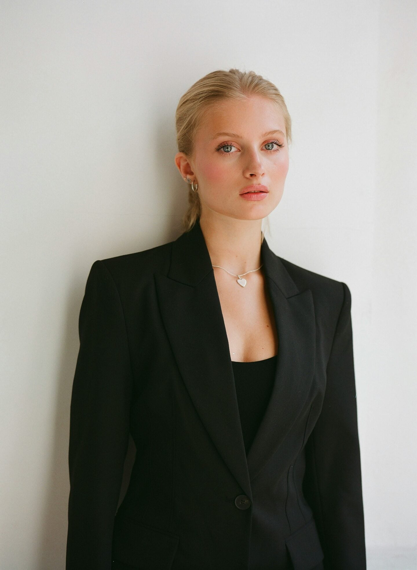 Professional young woman in a tailored black blazer and silver heart necklace, standing against a white wall with a confident and polished expression. Her blonde hair is pulled back neatly, creating a clean, modern business portrait.