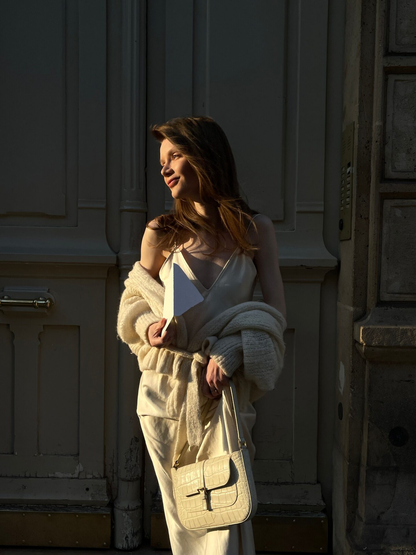 Young woman in a cream satin slip dress and oversized knit cardigan standing in warm golden-hour sunlight outside a historic building, holding a white envelope and a white crocodile-embossed shoulder bag. The soft lighting highlights her profile as she gazes into the distance, creating an elegant and timeless atmosphere.