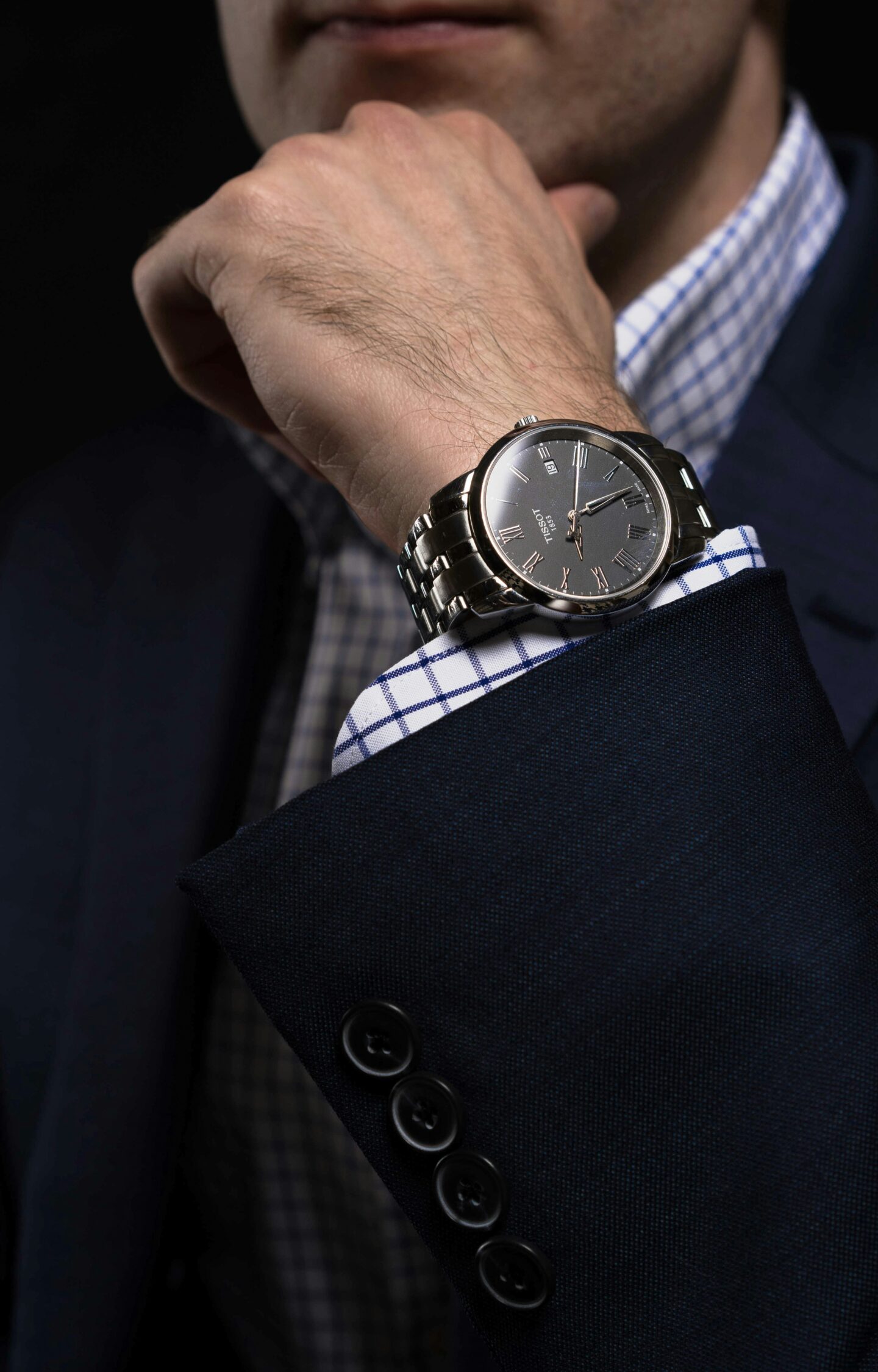 Close-up of a professionally dressed man wearing a dark suit jacket and checkered shirt, showcasing a silver wristwatch with a metal bracelet. The image highlights business attire, personal style, and professional appearance.