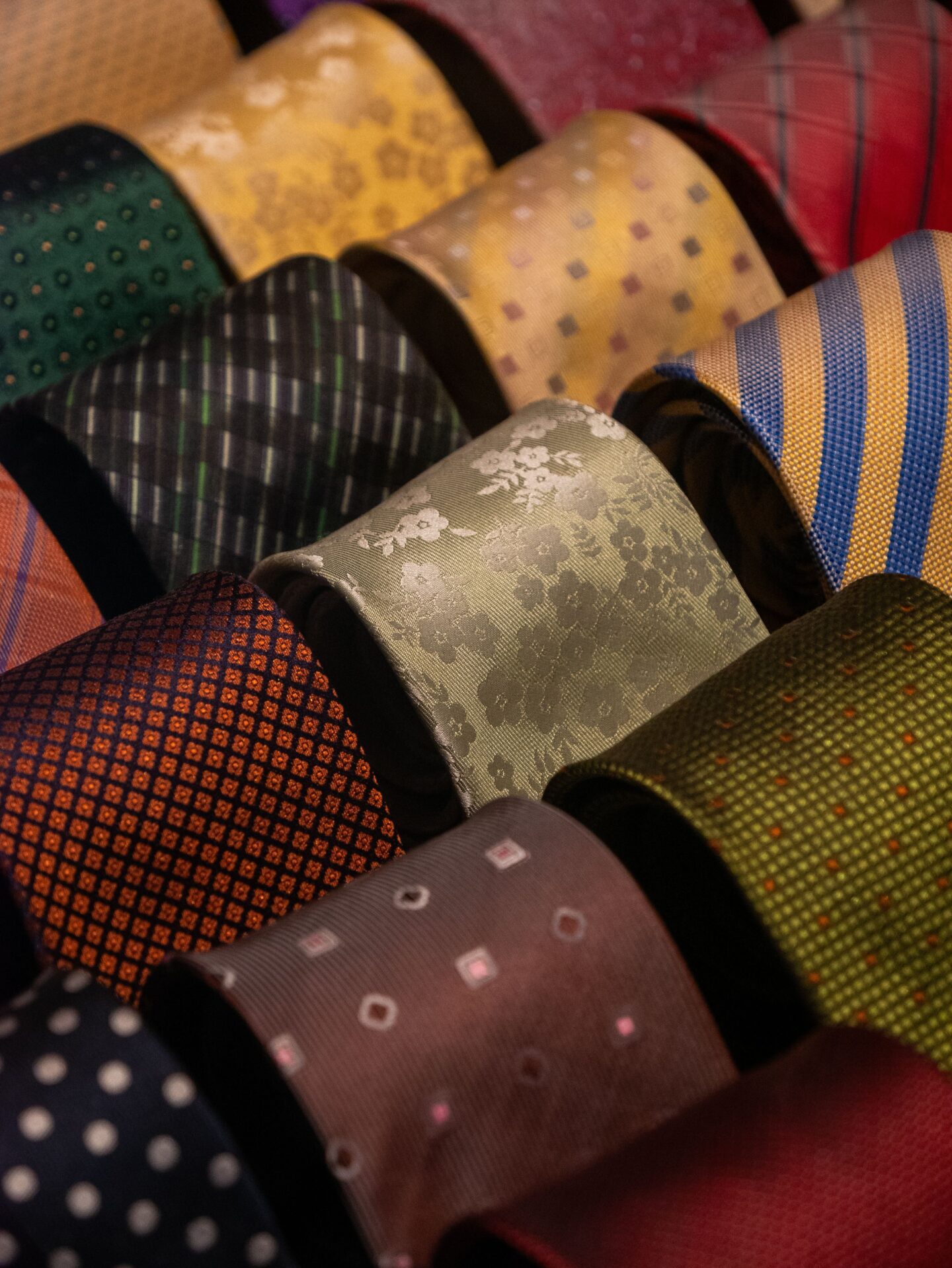 Collection of colorful patterned silk neckties displayed in rows, featuring stripes, florals, polka dots, checks, and geometric designs in shades of gold, blue, green, red, brown, and gray. The close-up view highlights the textures, intricate weaving, and variety of classic tie patterns.
