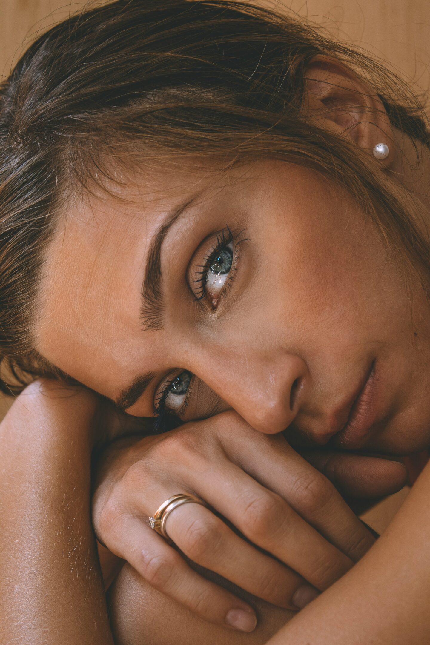 Close-up portrait of a woman resting her head on her arms, wearing a delicate gold ring and pearl stud earring. The image highlights natural beauty, soft makeup, and elegant jewellery in a warm, intimate setting.