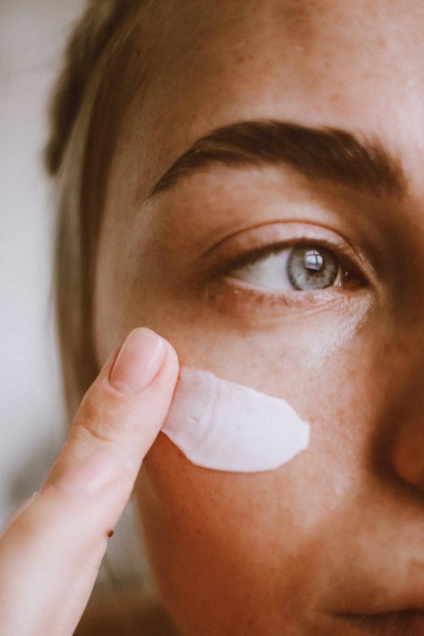 Close-up of a person applying white moisturiser or sunscreen to their cheek with a fingertip as part of a daily skincare routine.