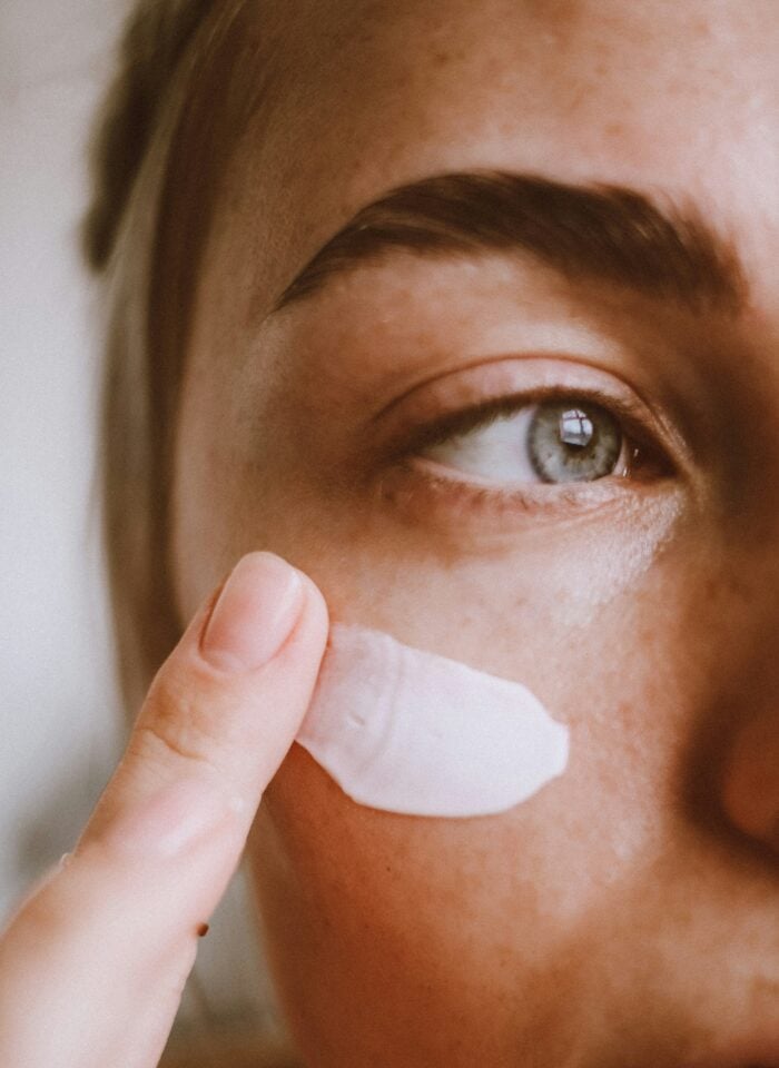 Close-up of a person applying white moisturiser or sunscreen to their cheek with a fingertip as part of a daily skincare routine.