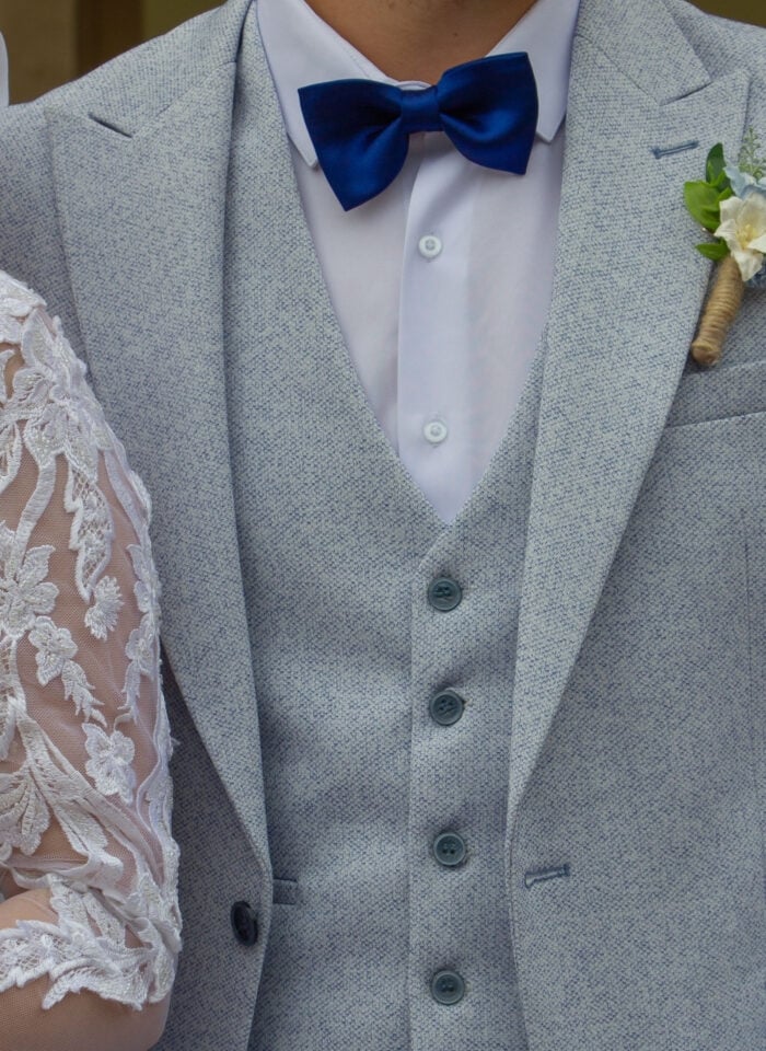 Groom wearing a light grey textured three-piece suit with a white shirt and royal blue satin bow tie, finished with a white floral boutonniere wrapped in twine. Partial view of the bride's white lace wedding dress is visible beside him.
