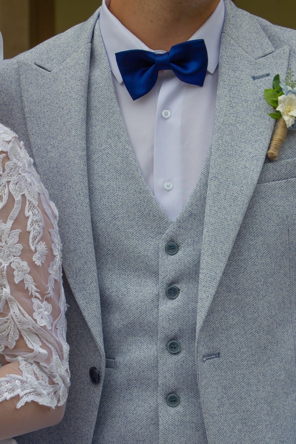 Groom wearing a light grey textured three-piece suit with a white shirt and royal blue satin bow tie, finished with a white floral boutonniere wrapped in twine. Partial view of the bride's white lace wedding dress is visible beside him.
