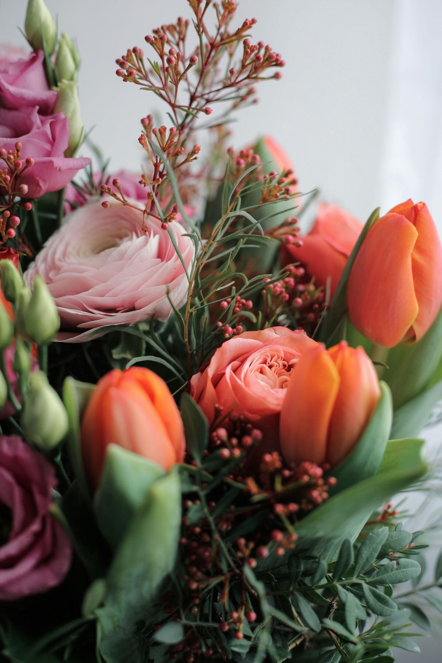Close-up of a fresh floral arrangement featuring peach tulips, pink roses, pale ranunculus, lisianthus buds, and greenery in soft natural light.