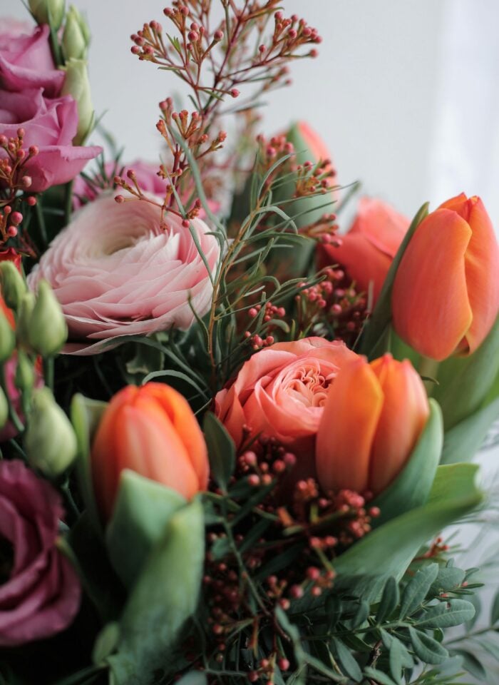Close-up of a fresh floral arrangement featuring peach tulips, pink roses, pale ranunculus, lisianthus buds, and greenery in soft natural light.