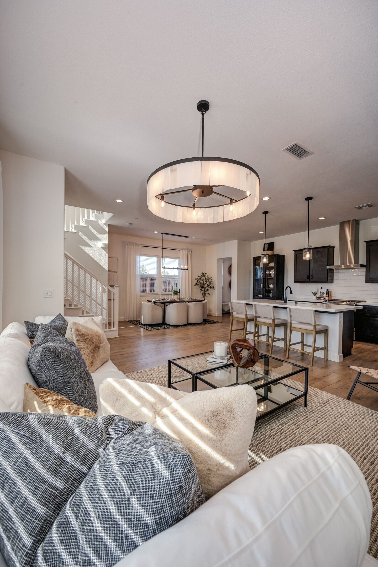 Bright open-plan living room with a white sofa, glass coffee table, modern kitchen island, dining area, wooden flooring, staircase, and large circular pendant light in a contemporary home.
