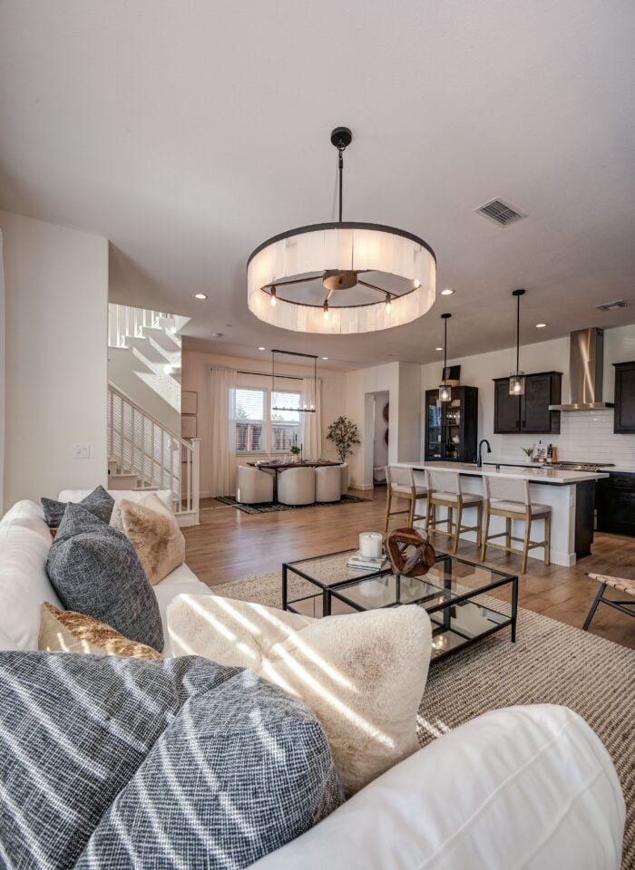 Bright open-plan living room with a white sofa, glass coffee table, modern kitchen island, dining area, wooden flooring, staircase, and large circular pendant light in a contemporary home.