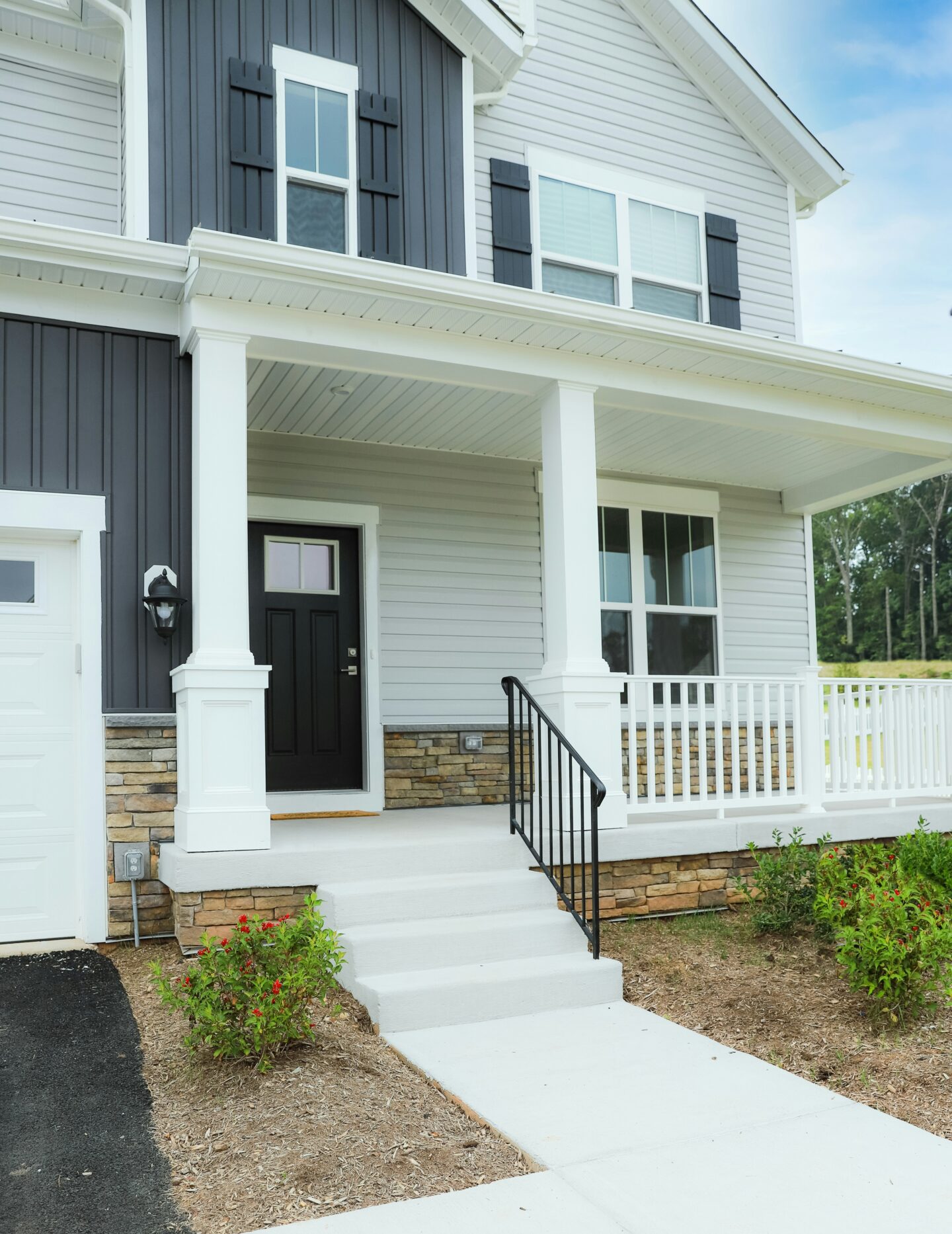 A modern two-story house with a covered front porch, white columns, and a black front door. The exterior combines light gray horizontal siding with dark charcoal vertical panels and matching shutters, accented by stacked stone along the lower walls. White railings border the porch, black metal handrails line the concrete steps, and small flowering shrubs sit in mulched garden beds beside the front walkway. A portion of the attached garage and driveway is visible on the left, with trees and a blue sky in the background.