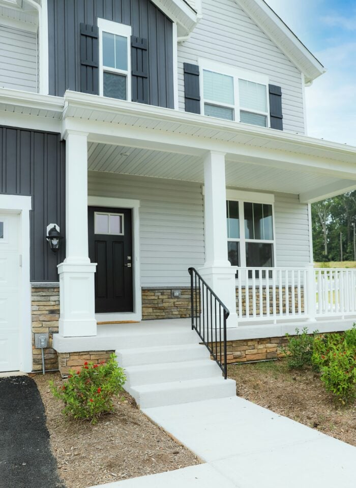 A modern two-story house with a covered front porch, white columns, and a black front door. The exterior combines light gray horizontal siding with dark charcoal vertical panels and matching shutters, accented by stacked stone along the lower walls. White railings border the porch, black metal handrails line the concrete steps, and small flowering shrubs sit in mulched garden beds beside the front walkway. A portion of the attached garage and driveway is visible on the left, with trees and a blue sky in the background.