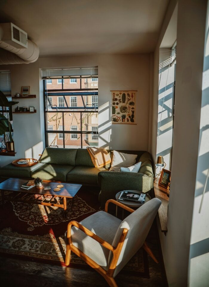 Cozy small living room with a green sofa, wooden coffee table, accent chair, large windows, and warm natural sunlight creating an open, airy feel.