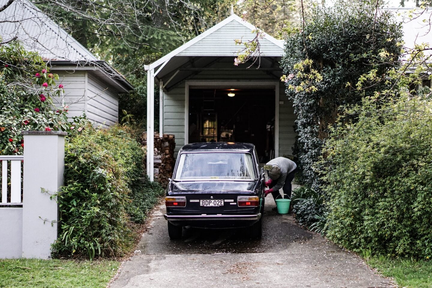 A person washes a black classic car parked in a narrow residential driveway outside an open garage. The driveway is lined with dense green hedges and flowering shrubs, with a bucket of water beside the car, creating a quiet suburban scene focused on classic car care and maintenance.
