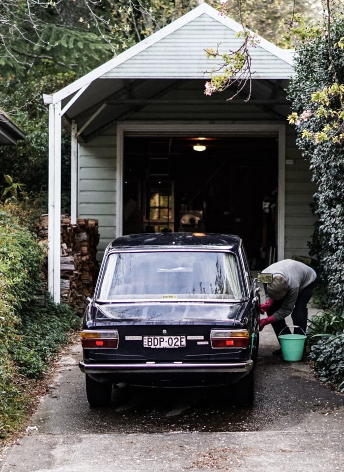 A person washes a black classic car parked in a narrow residential driveway outside an open garage. The driveway is lined with dense green hedges and flowering shrubs, with a bucket of water beside the car, creating a quiet suburban scene focused on classic car care and maintenance.