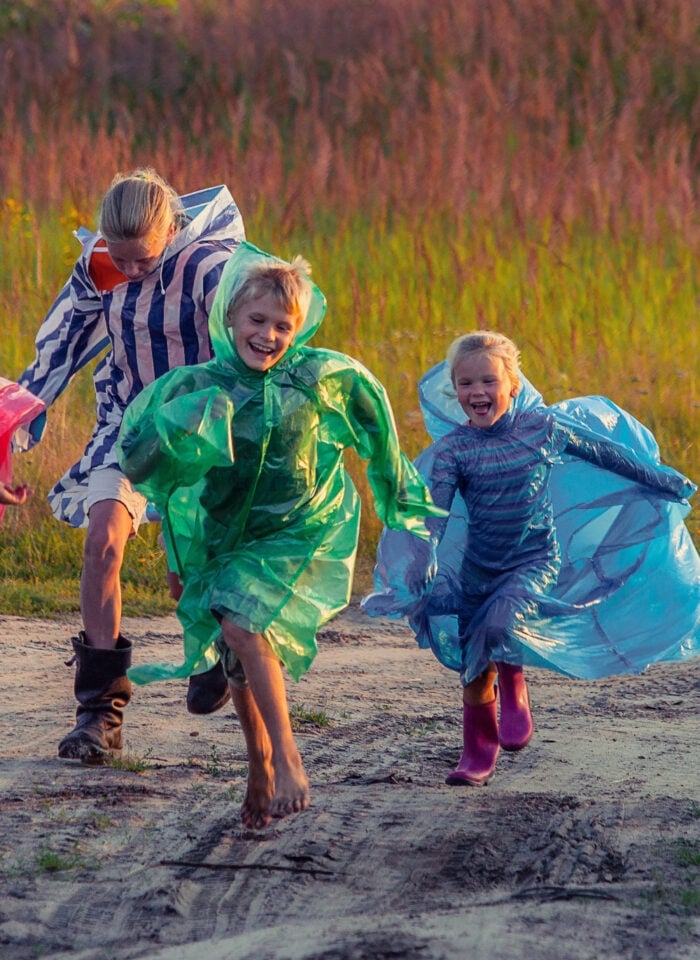 Five children wearing colorful rain ponchos and rubber boots run along a dirt path through a grassy field. Smiling and laughing, they play outdoors despite the wet weather, with one child barefoot and others in bright pink, green, blue, and striped rain gear. Warm evening sunlight illuminates the scene, creating a cheerful atmosphere surrounded by tall grass and trees.