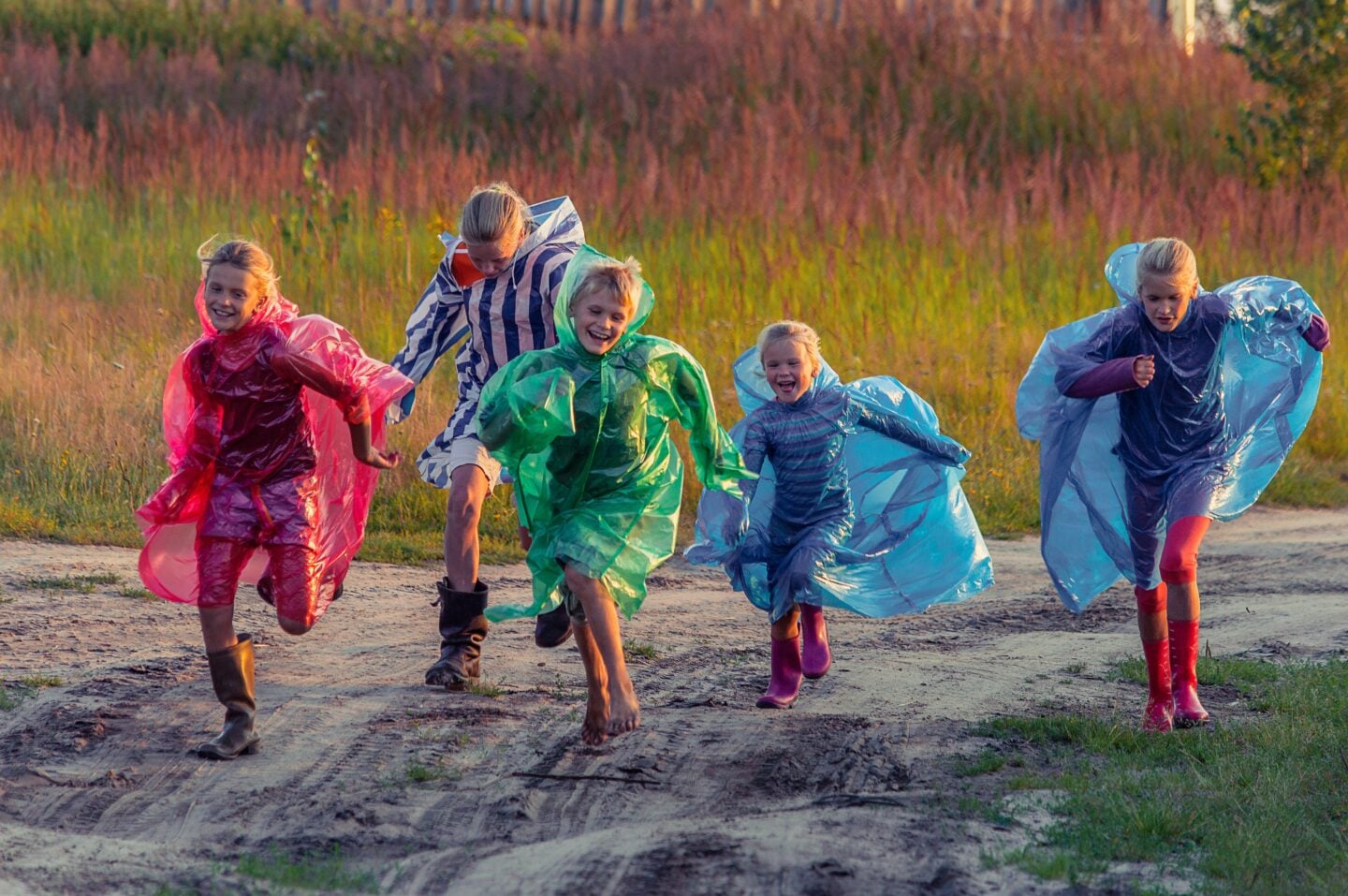 Five children wearing colorful rain ponchos and rubber boots run along a dirt path through a grassy field. Smiling and laughing, they play outdoors despite the wet weather, with one child barefoot and others in bright pink, green, blue, and striped rain gear. Warm evening sunlight illuminates the scene, creating a cheerful atmosphere surrounded by tall grass and trees.