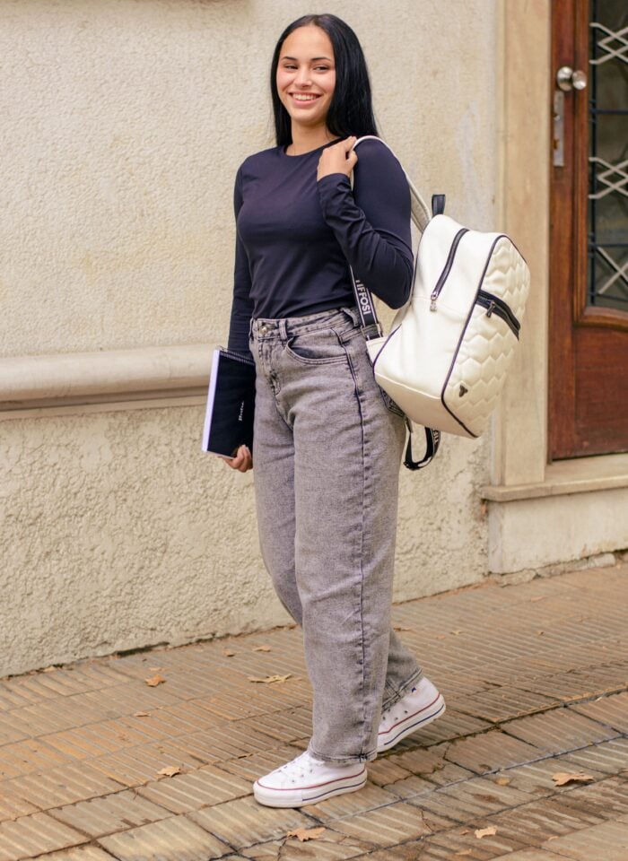A smiling girl walks along a paved pathway outside a building, carrying a white backpack over one shoulder and a notebook in one hand. She is dressed casually in a black long-sleeve top, gray jeans, and white sneakers, with a wooden doorway and textured exterior wall in the background.