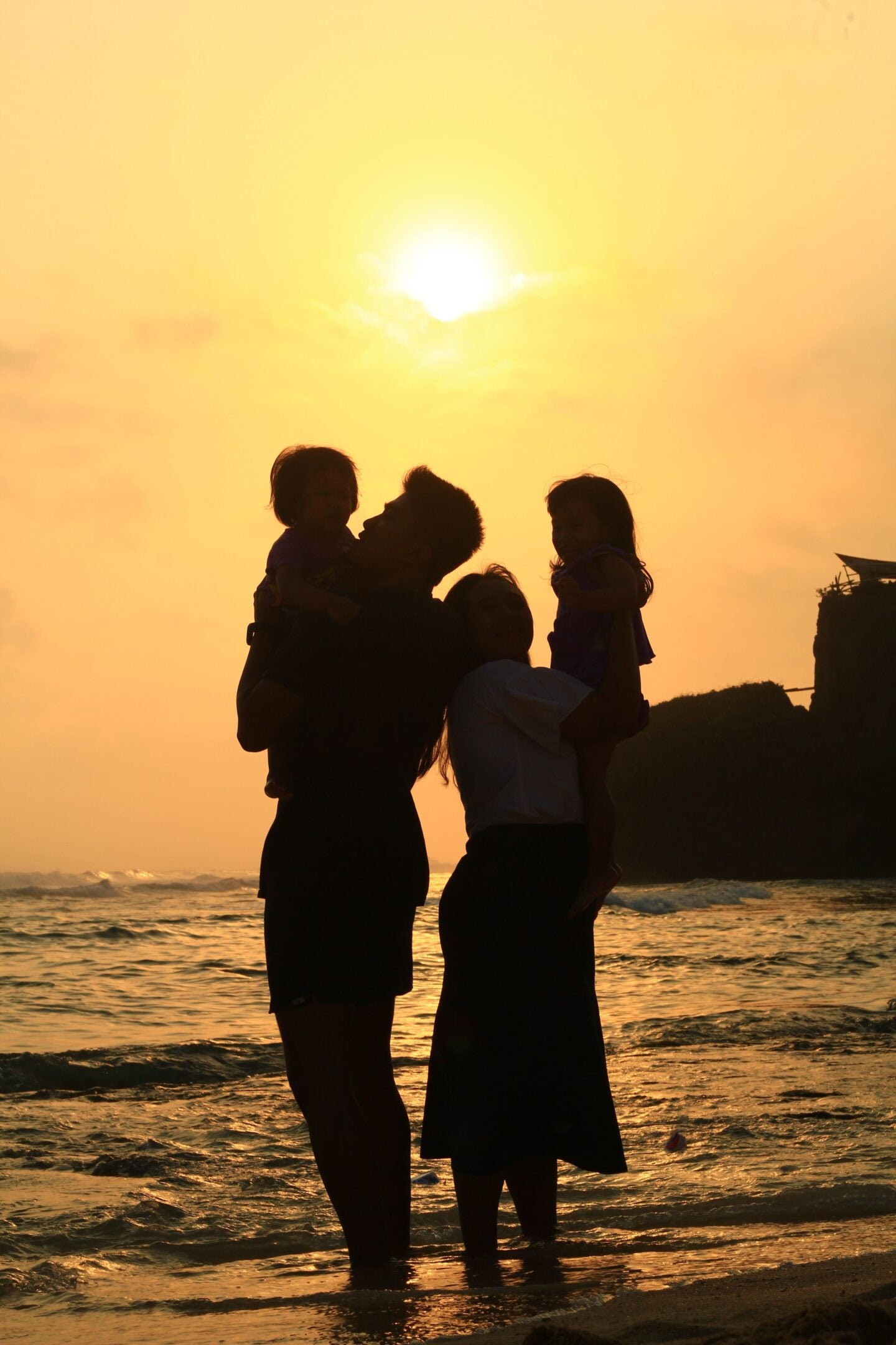 Silhouettes of two parents standing in shallow ocean water at sunset, each holding a young child. The family faces one another against a golden sky, with gentle waves and a rocky coastline creating a warm, peaceful backdrop that symbolizes togetherness and life's important milestones.
