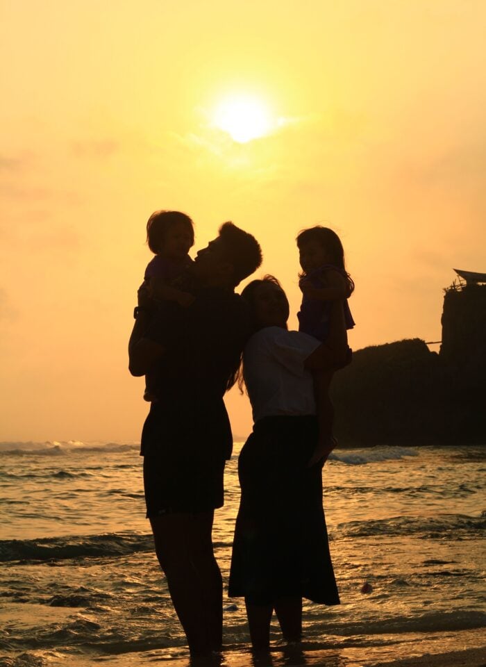 Silhouettes of two parents standing in shallow ocean water at sunset, each holding a young child. The family faces one another against a golden sky, with gentle waves and a rocky coastline creating a warm, peaceful backdrop that symbolizes togetherness and life's important milestones.