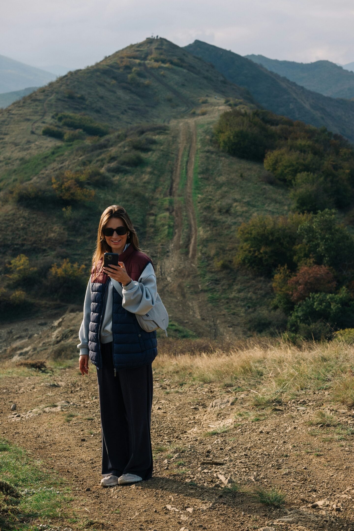 A person wearing sunglasses, a quilted puffer vest over a light hoodie, dark wide-leg trousers, and white trainers stands on a dirt hiking trail while looking at a smartphone. Behind them, a steep ridgeline with twin dirt tracks climbs toward a hilltop viewpoint, surrounded by rolling green and brown mountains under a cloudy sky.
