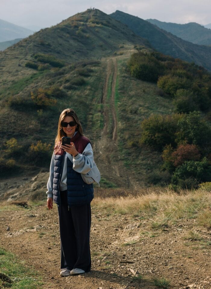 A person wearing sunglasses, a quilted puffer vest over a light hoodie, dark wide-leg trousers, and white trainers stands on a dirt hiking trail while looking at a smartphone. Behind them, a steep ridgeline with twin dirt tracks climbs toward a hilltop viewpoint, surrounded by rolling green and brown mountains under a cloudy sky.