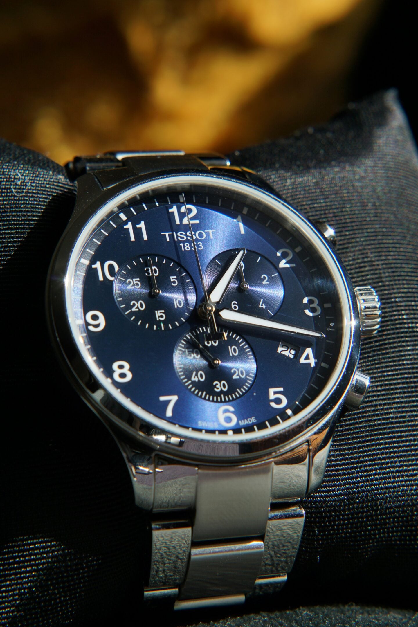 Close-up of a stainless steel Tissot chronograph watch with a blue dial resting on a dark fabric surface.