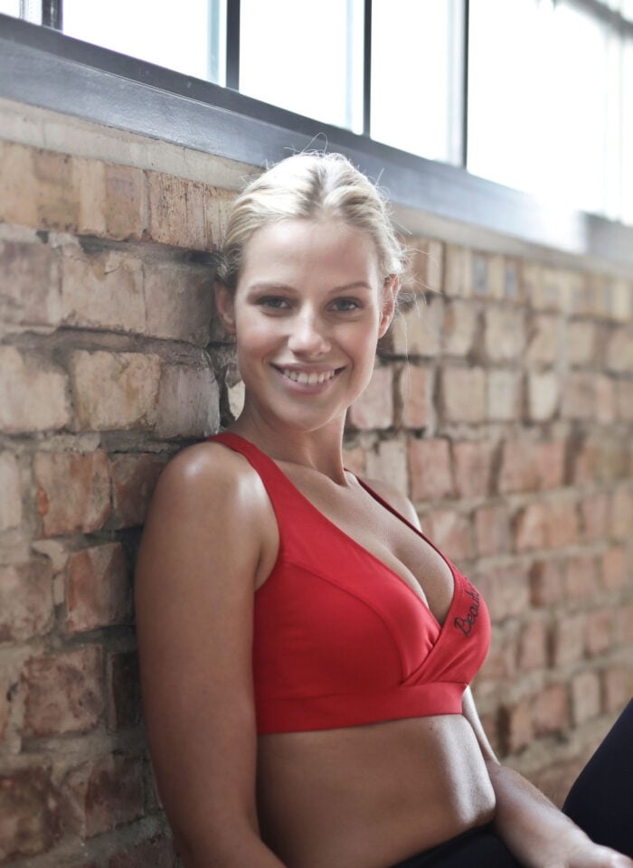Smiling woman in a red sports bra and black leggings sitting against an exposed brick wall, holding pink dumbbells after a workout in a bright gym setting.
