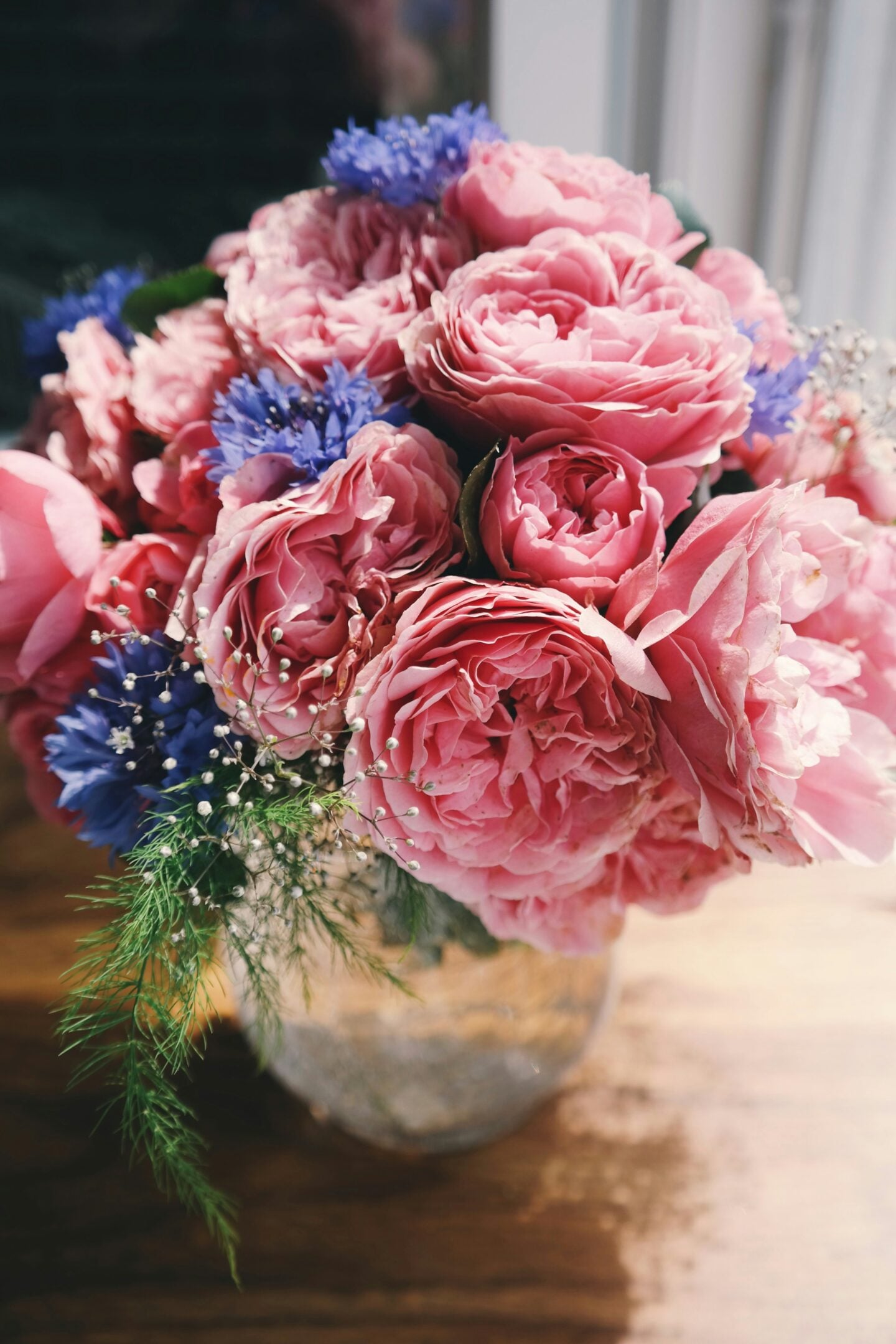Elegant bouquet of pink garden roses, blue cornflowers, baby's breath, and fresh greenery arranged in a rustic glass vase on a wooden table.