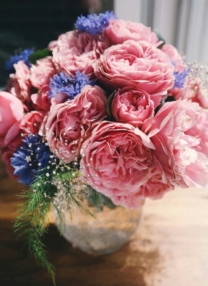 Elegant bouquet of pink garden roses, blue cornflowers, baby's breath, and fresh greenery arranged in a rustic glass vase on a wooden table.